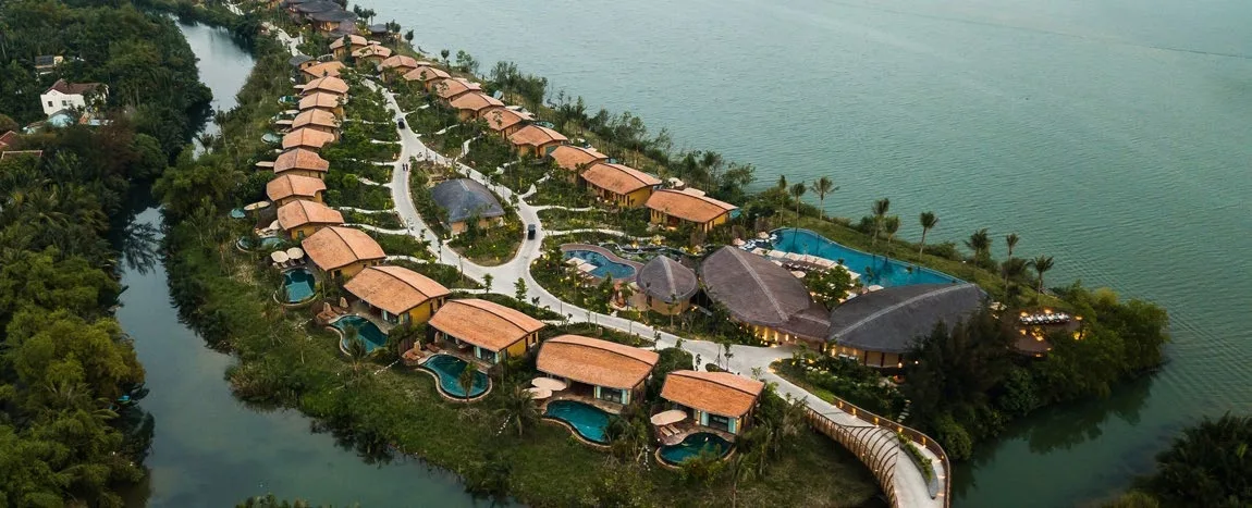 Aerial view of Namia River Retreat, a line of villas with private pools on an island in the Thu Bon River